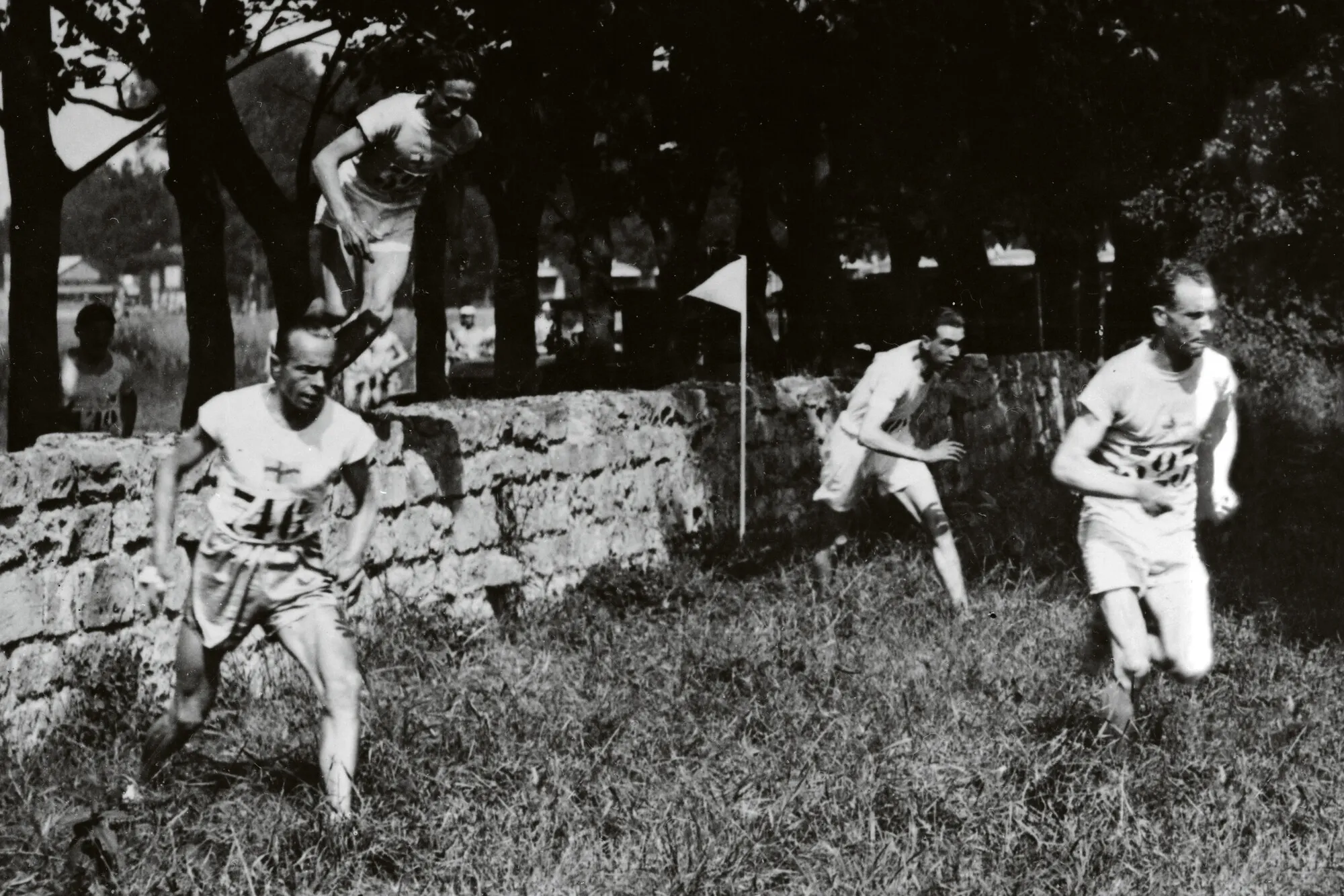 Paavo Nurmi johtaa läkähdyttävässä helteessä käytyä maastojuoksua. Edvin Wide näkyy vasemmalla ja Ville Ritola ylittää paraikaa kiviaitaa.