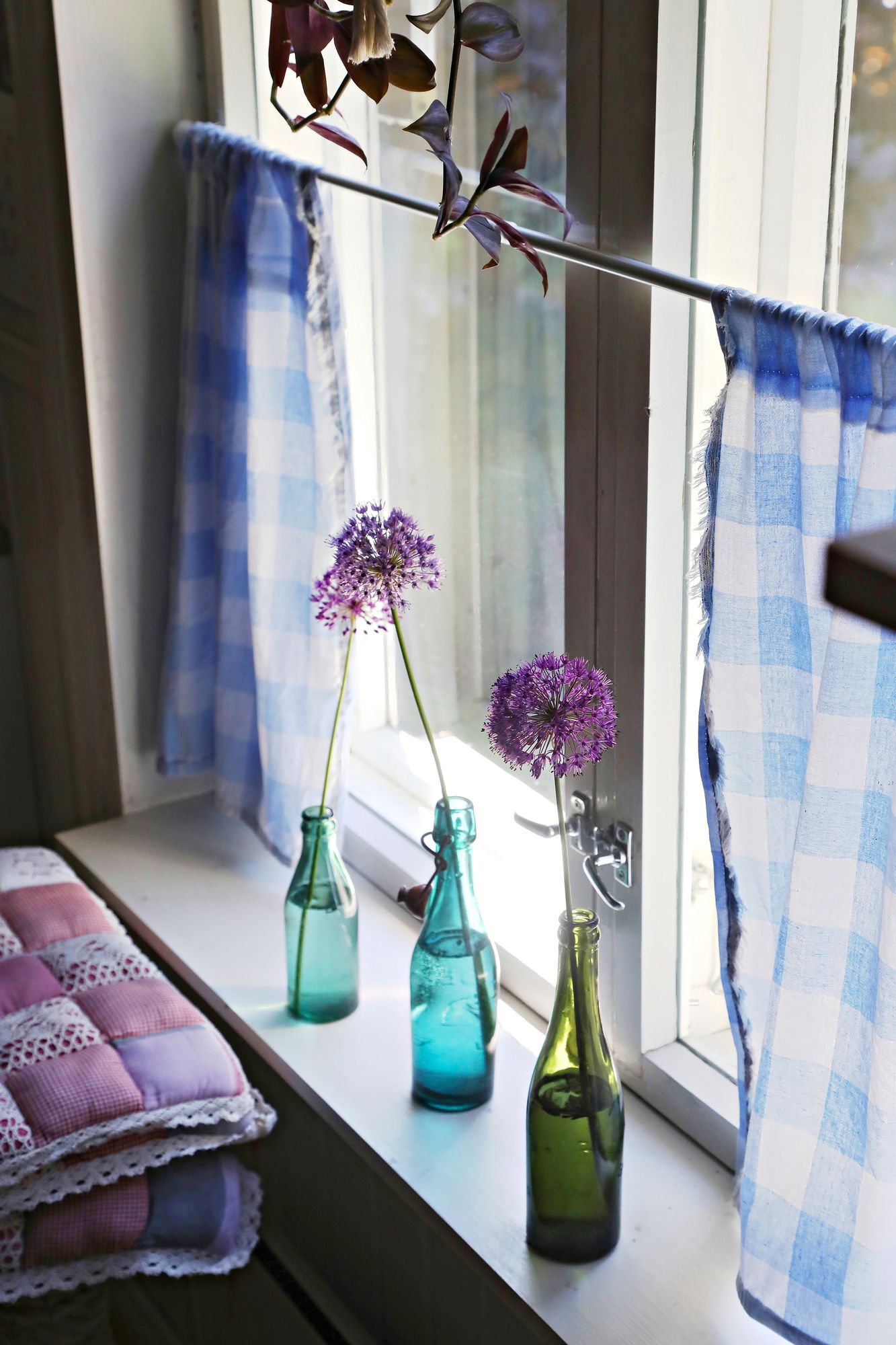 Vanhat lasi­pullot loistavat ikkunalla. Jätti­laukan kukat ovat omalta pihalta. © Mikko Kaaresmaa
